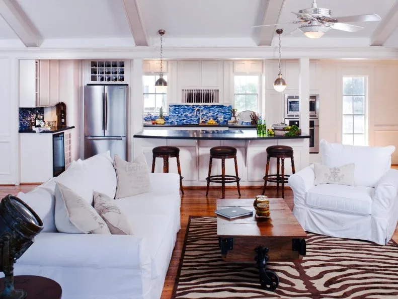 White sofas facing a wooden coffee table and striped rug in front of a pale kitchen with a blue patterned backsplash.