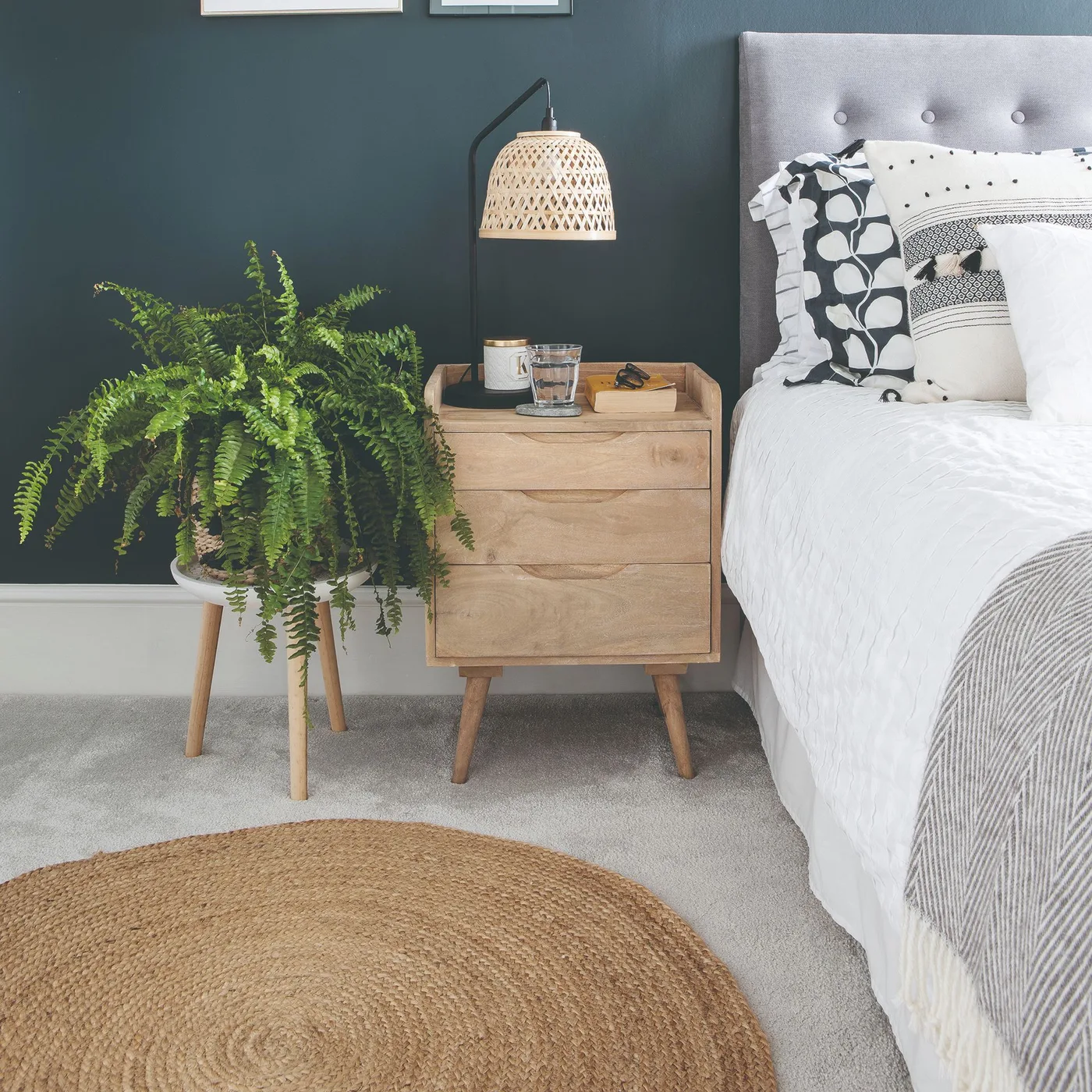 Dark blue-gray bedroom wall with a gray tufted headboard, wooden bedside table, woven pendant, fern, and white bedding.
