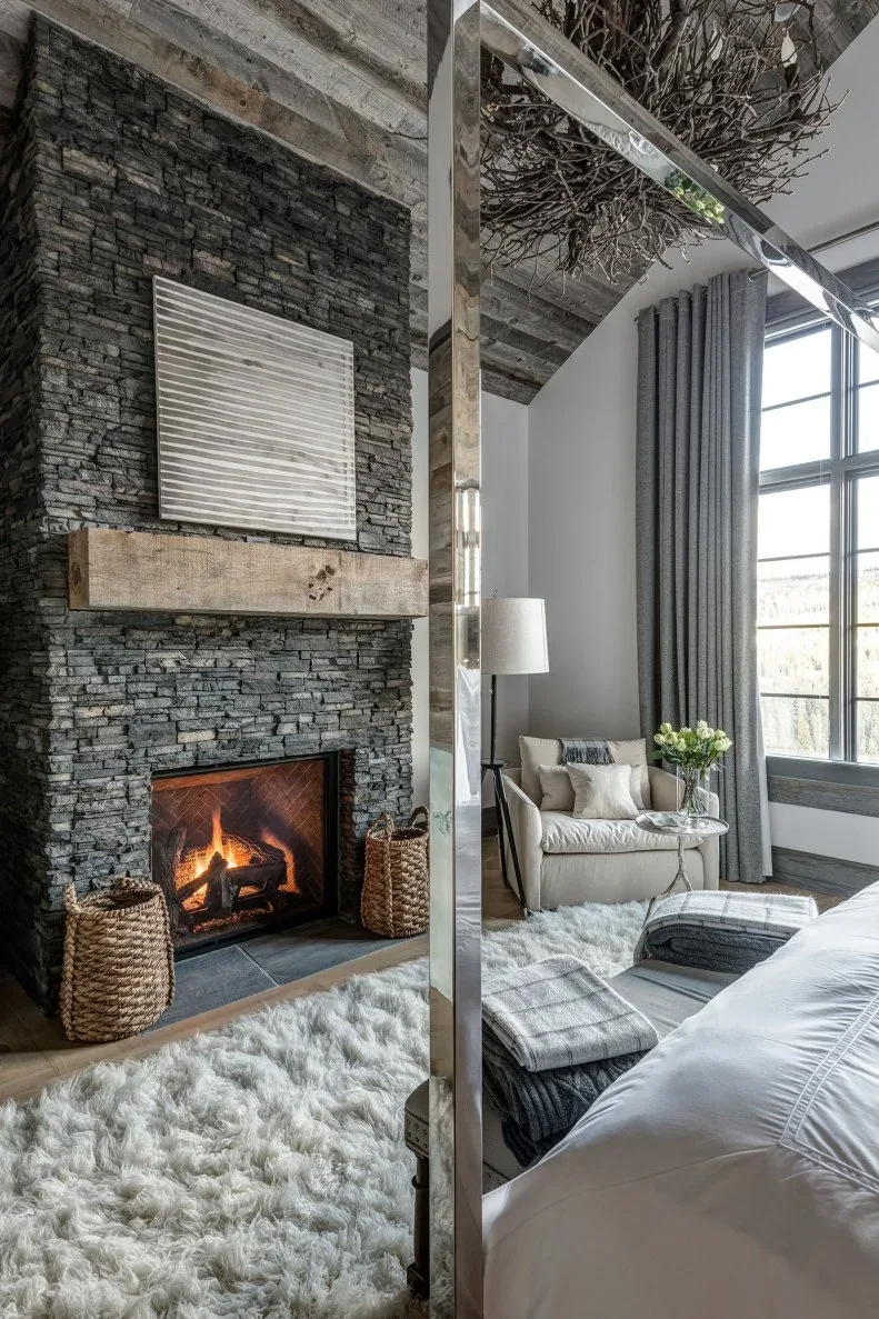 Tall dark stacked-stone fireplace in bedroom with two woven baskets, light chair and pale rug