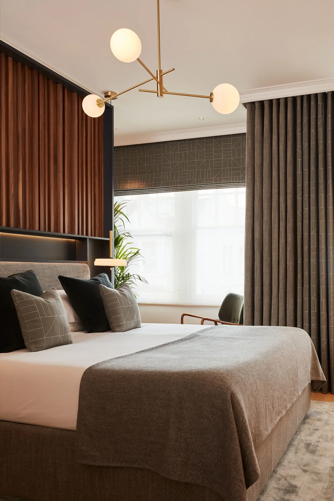 A bedroom combines vertical wood details behind dark pillows, pale bedding and a gray throw beneath a three-globe pendant beside tall gray curtains