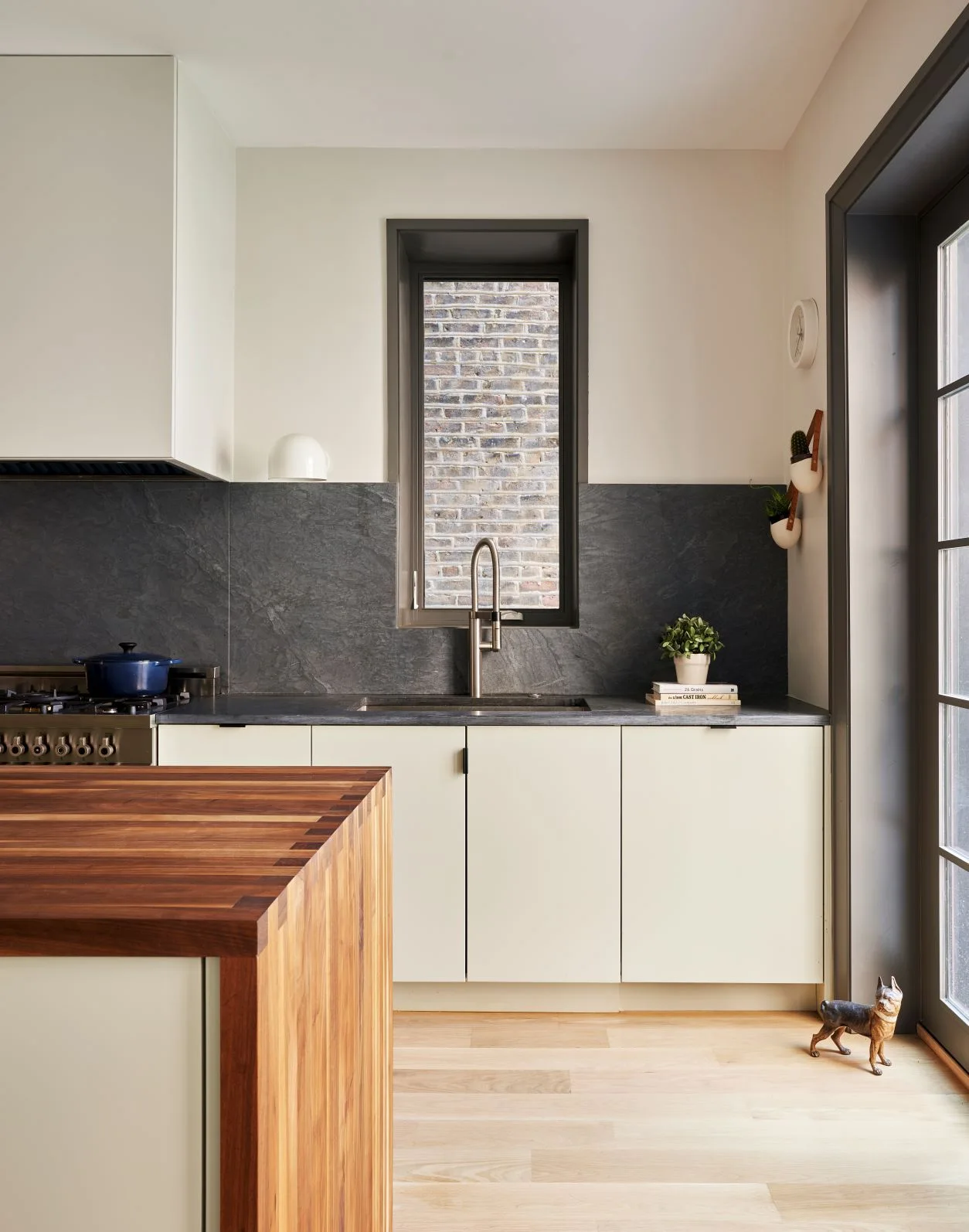 Light beige flat-front kitchen cabinets sit beneath a dark backsplash beside a narrow window, wooden island and black-framed doors
