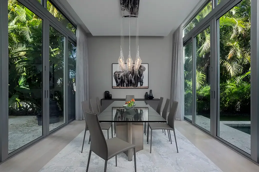 A clear rectangular dining table and gray chairs sit between tall dark-framed windows beside pale curtains, a light rug and hanging clear lights