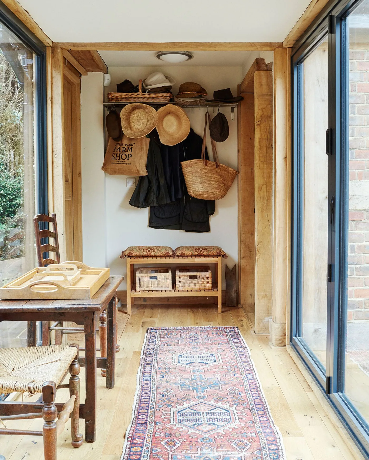 Glazed country passage with wooden framing, hanging hats and coats, a small bench with baskets, narrow runner and a table at the side.