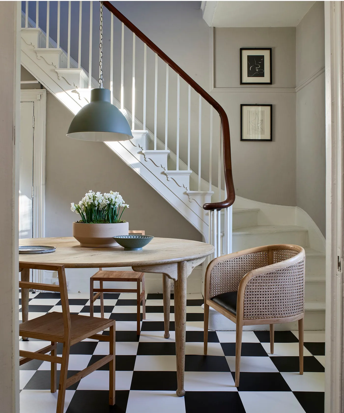 Dining space beside a traditional white staircase with a checkerboard floor, round wooden table and woven-back chair.