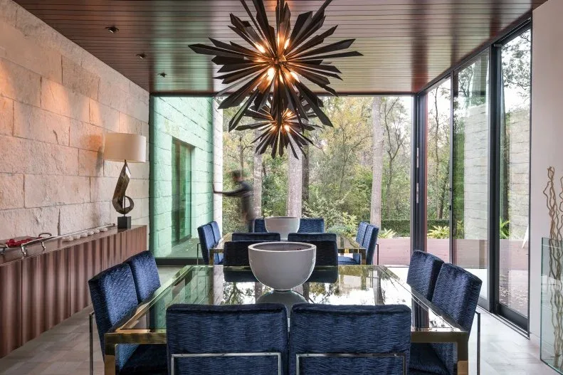Dining room with a pale block-pattern textured wall, long brown sideboard, glass table, blue chairs, dark ceiling, starburst lights, and full-height windows.
