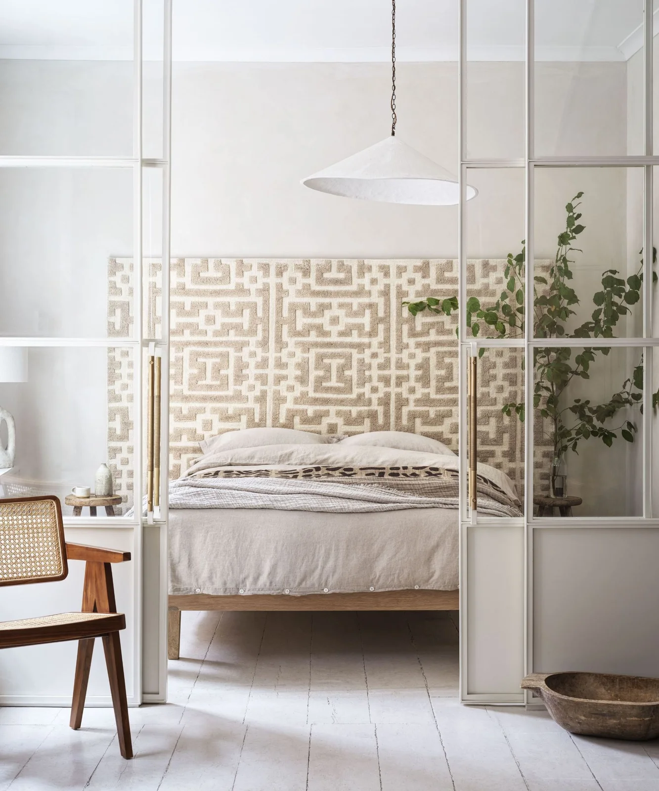 A pale bed with a patterned headboard sits behind a light grid-framed glazed partition near a wooden chair and large white pendant