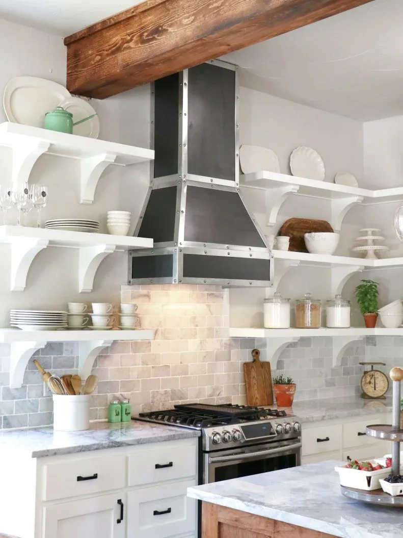 White framed kitchen cabinets with pale veined rectangular backsplash tiles, a dark hood and open shelves supported by white brackets.