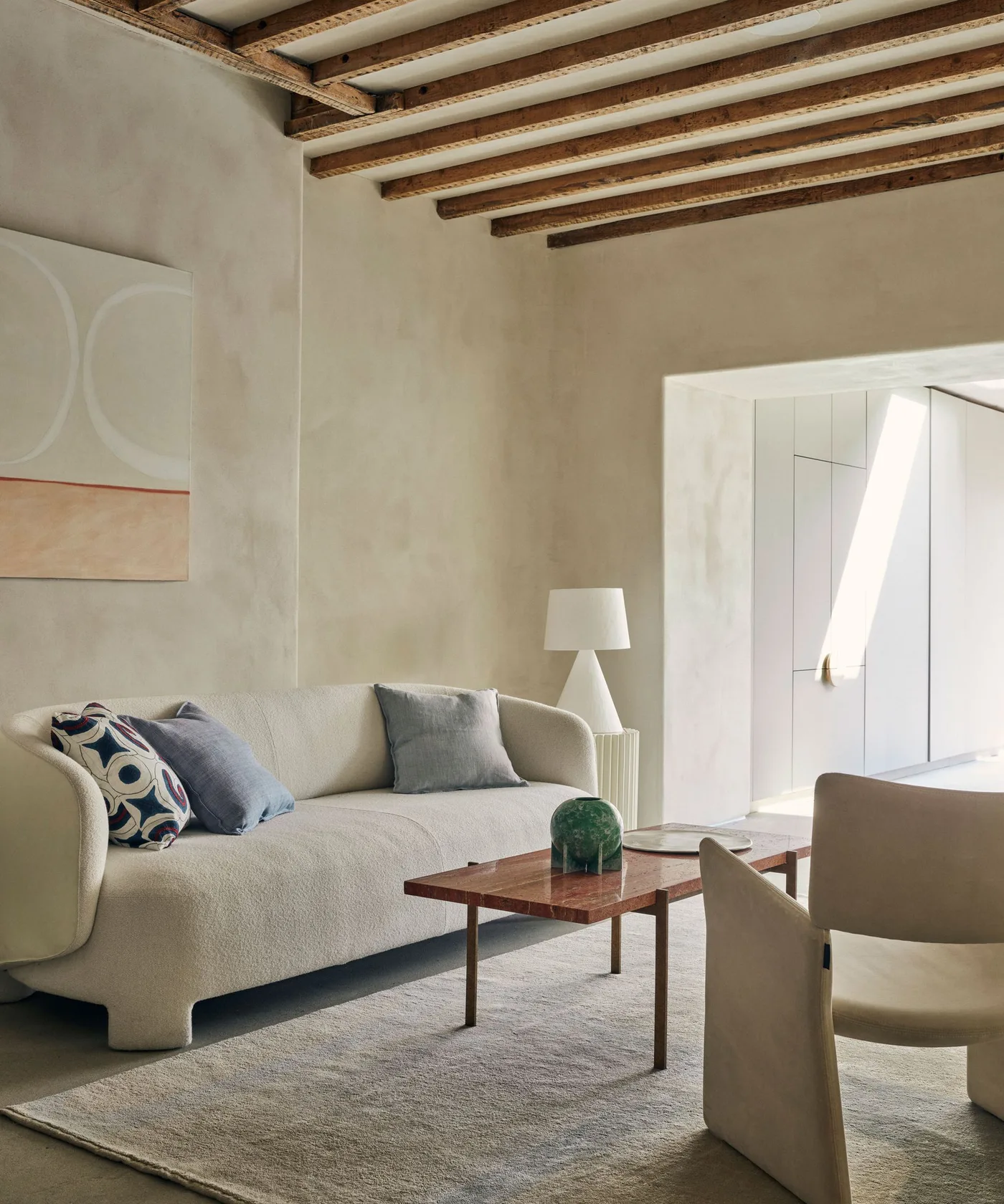 A pale curved sofa and light armchair face a narrow reddish wood table beneath exposed ceiling beams and beside a pale rug
