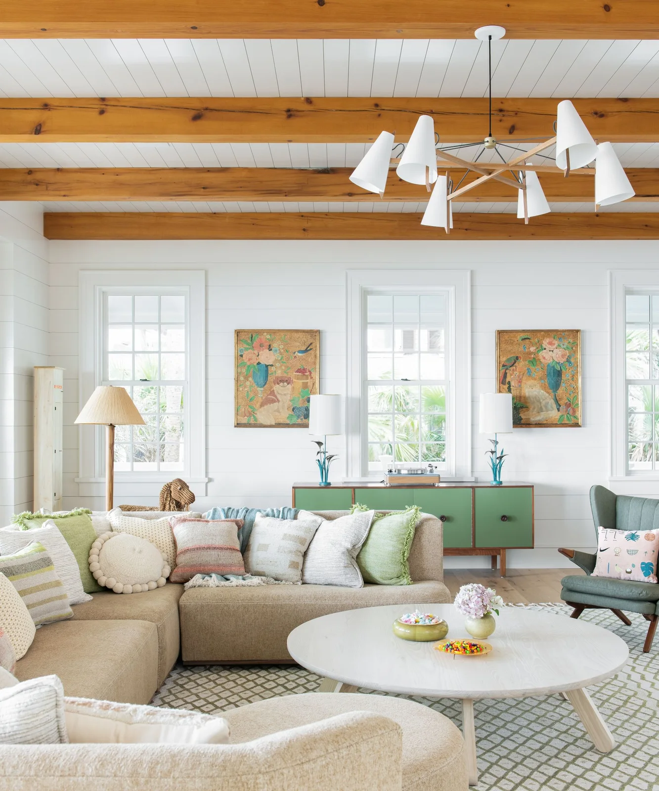 A green-fronted sideboard and green chair sit behind pale sectional seating and an oval coffee table beneath wooden ceiling beams