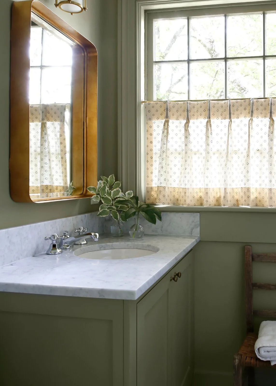 Sage green wash area with pale veined counter, warm-toned mirror and patterned cafe curtain