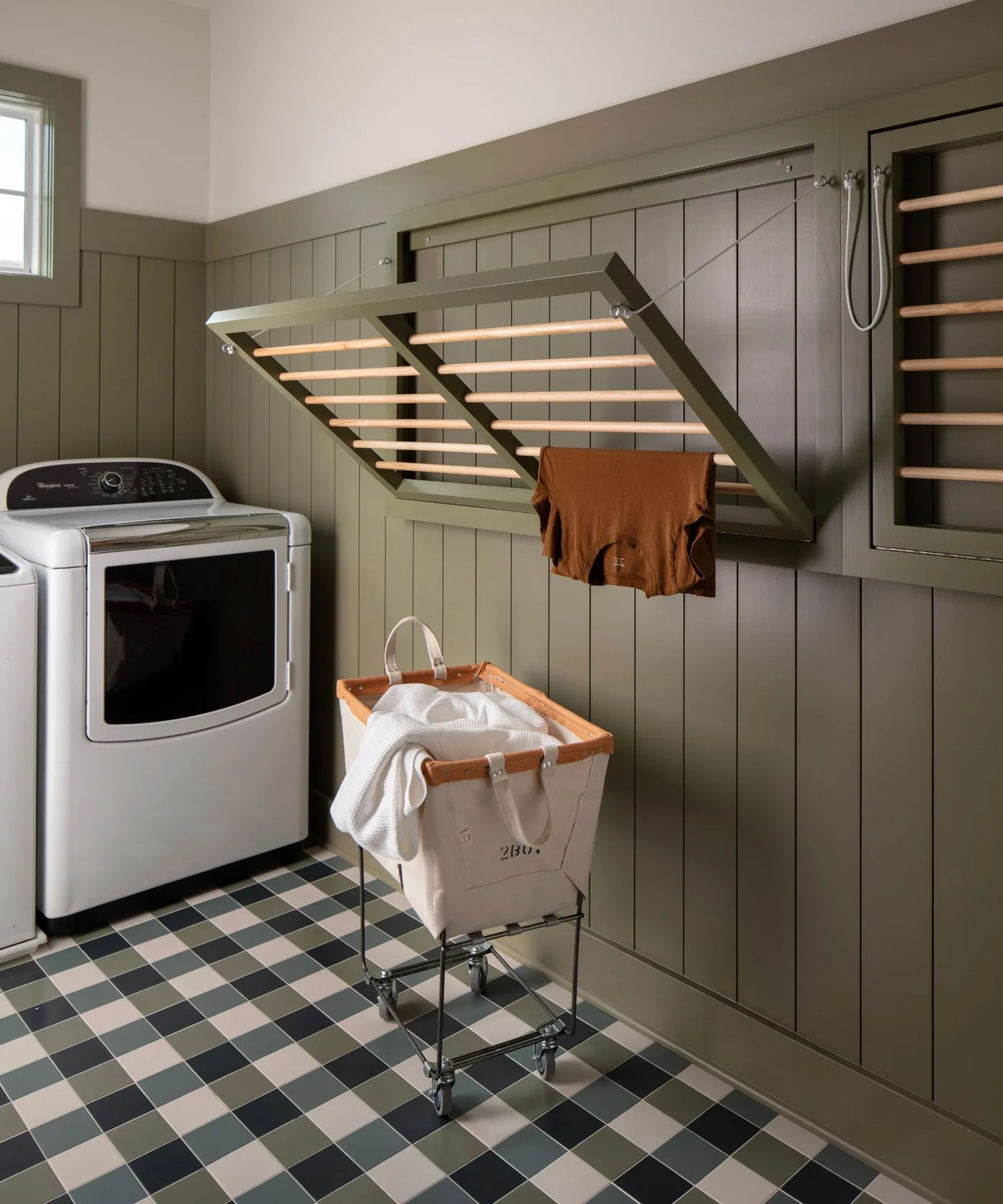 Green paneled laundry wall with hinged slatted drying rack, checked floor, top-loading machine and wheeled hamper