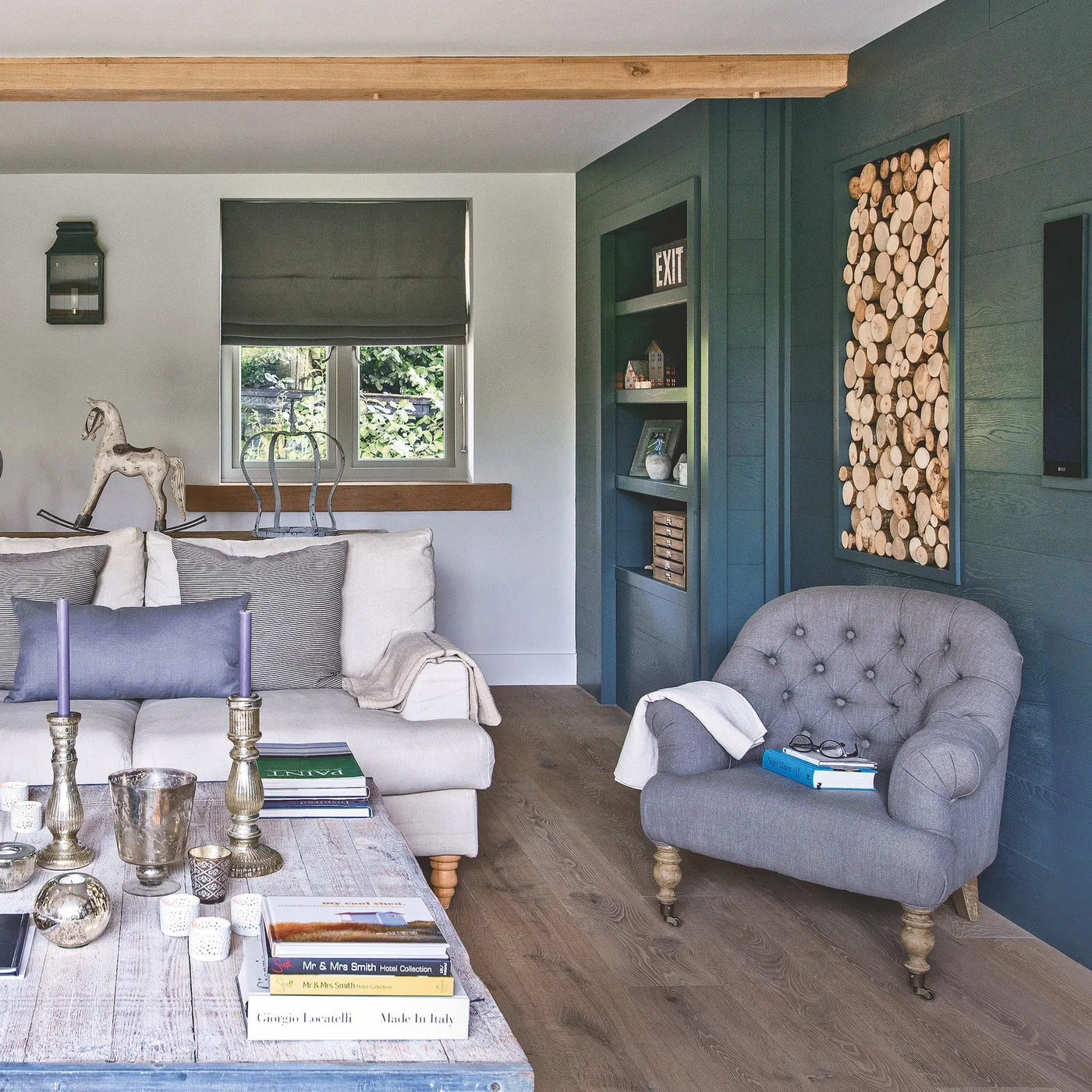 A pale sofa and rounded gray chair sit beside deep green walls, wooden trim and a framed display of log ends