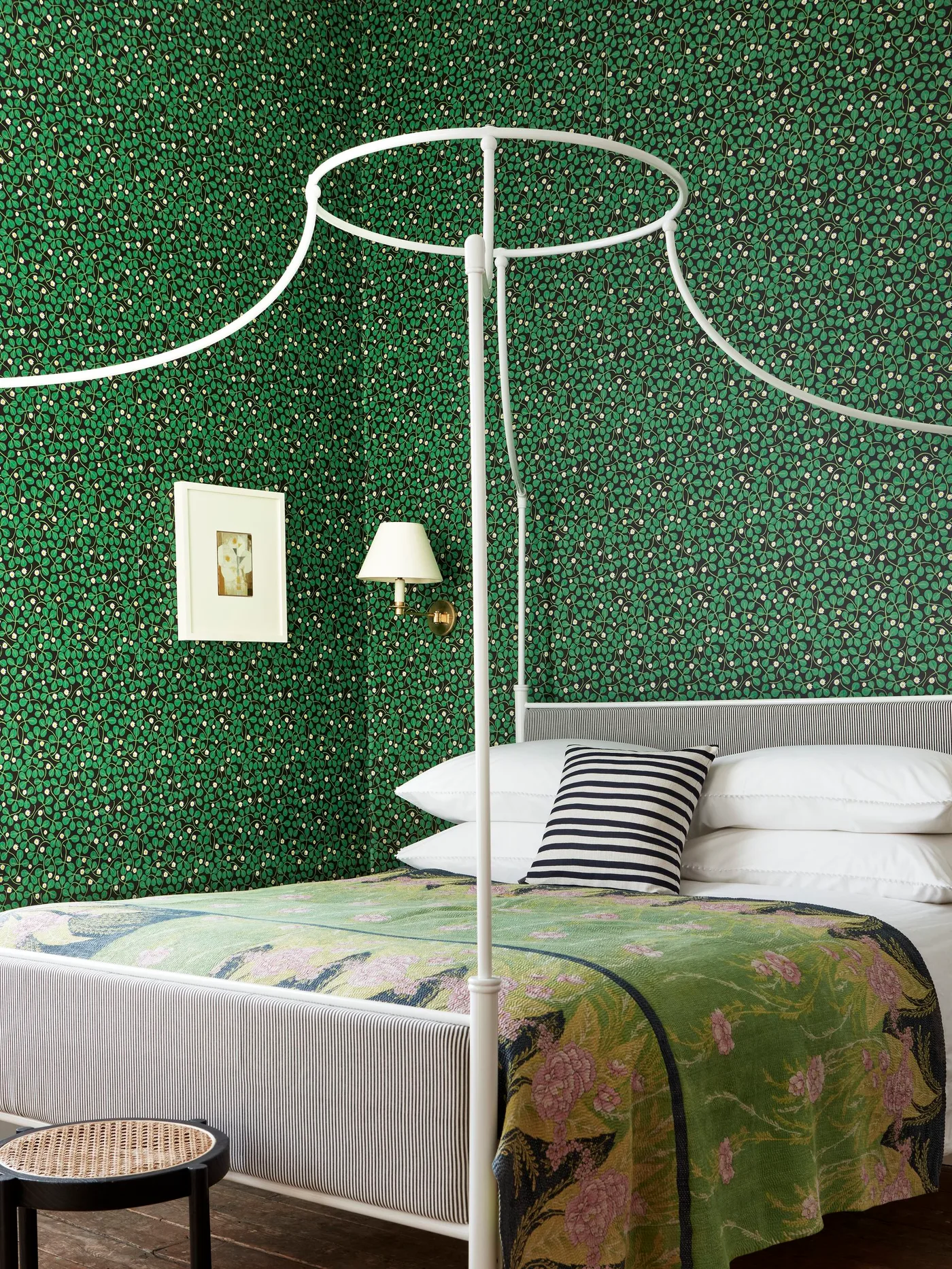 Bedroom with dense green patterned wallpaper, white metal canopy bed, floral green bedspread, striped cushion, and small wall light.