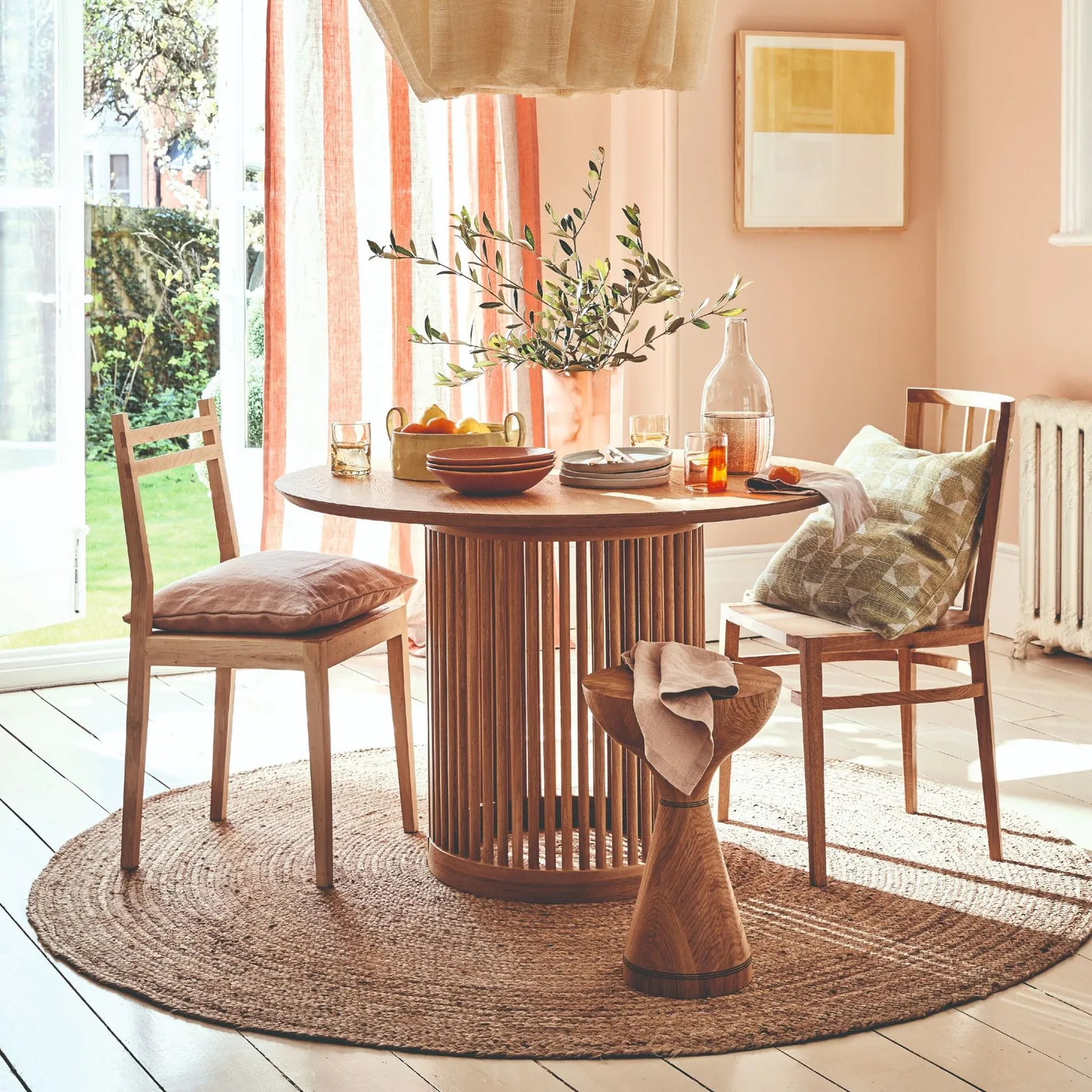 A round wooden table with a slatted base stands on a circular woven-looking rug beside wooden chairs, peach walls and warm striped curtains