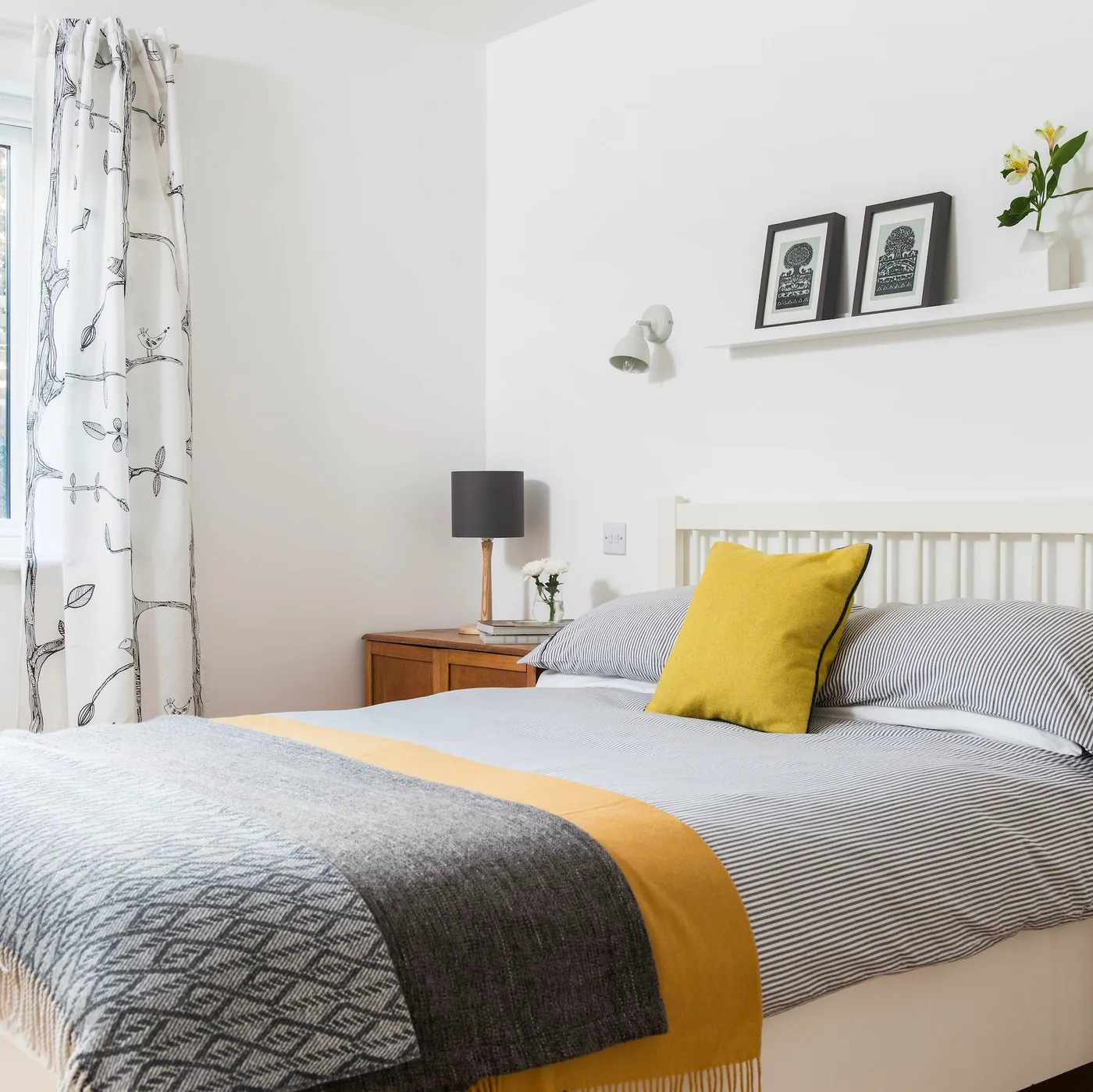 White bedroom with a pale spindle bed, striped bedding, mustard cushions, a wood bedside table and framed pictures on a ledge
