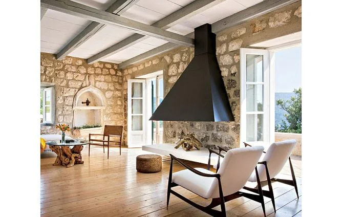 A broad dark fireplace hood stands against rough stone walls between pale upholstered wooden chairs and open windows