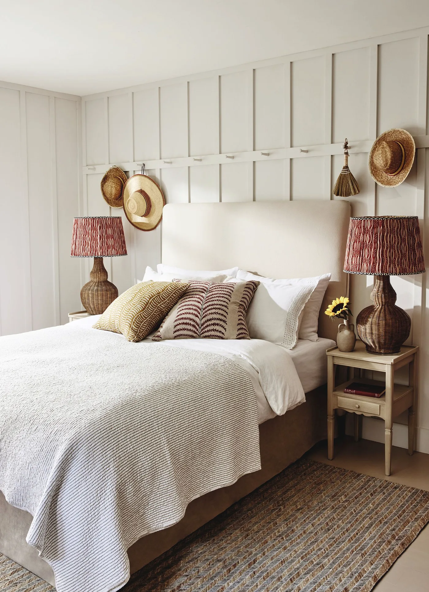 Bedroom with pale wall paneling, white upholstered headboard, woven bedside lamps with patterned red shades, hats on a rail and layered cushions.