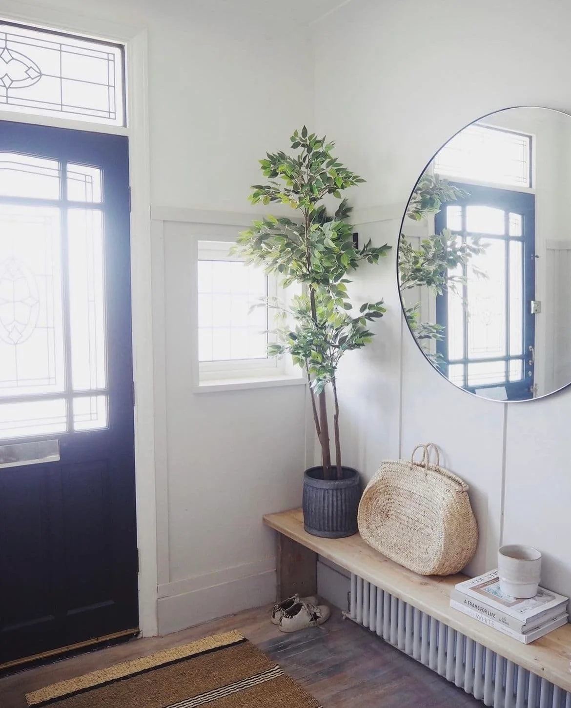 A wooden ledge above a white radiator holds a leafy potted plant, woven bag and books beneath a round mirror beside a dark glazed front door
