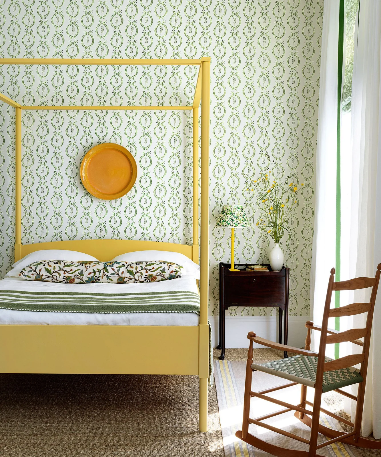 A yellow four-poster bed stands before green chain-pattern wallpaper with floral pillows, striped bedding and a yellow round wall decoration