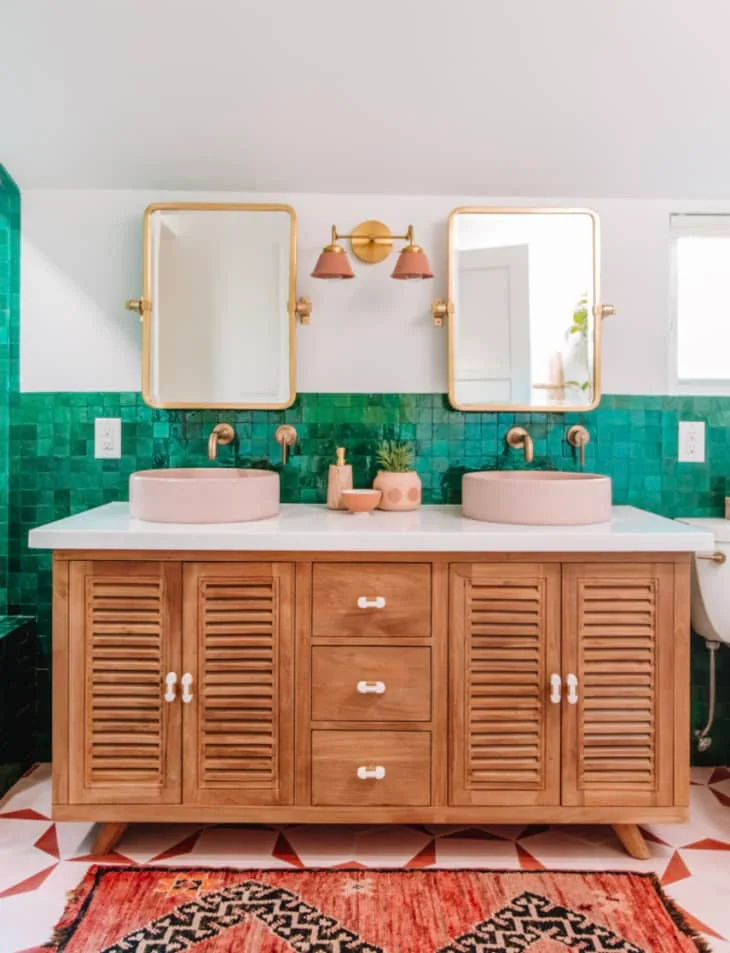 Pink vessel basins sit on a wooden vanity beneath two gold-toned mirrors beside emerald tiles and a patterned rug