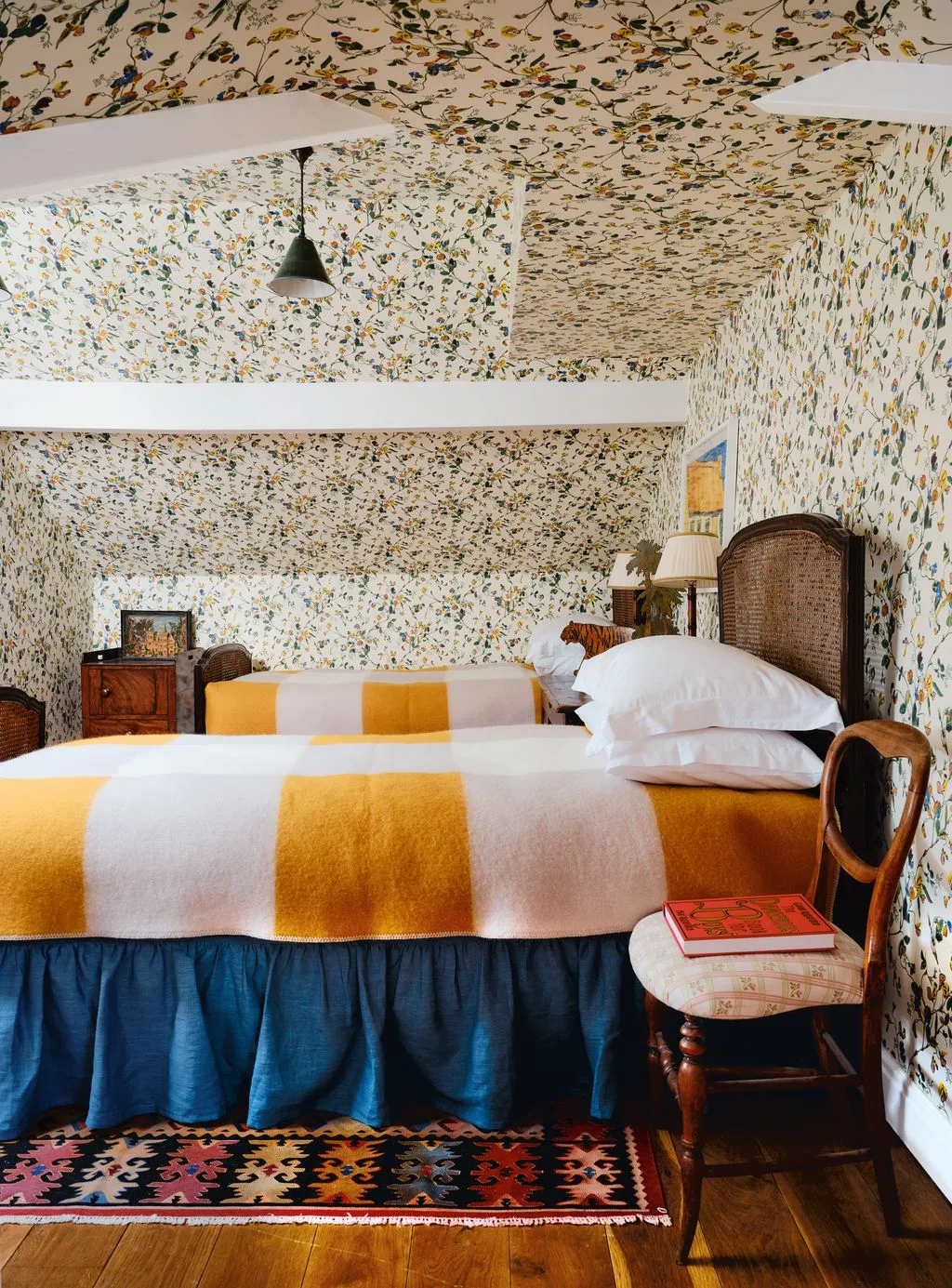 Twin attic bedroom with floral walls and ceiling, woven-panel headboards, yellow-and-white checked covers, blue bed skirts and white beams