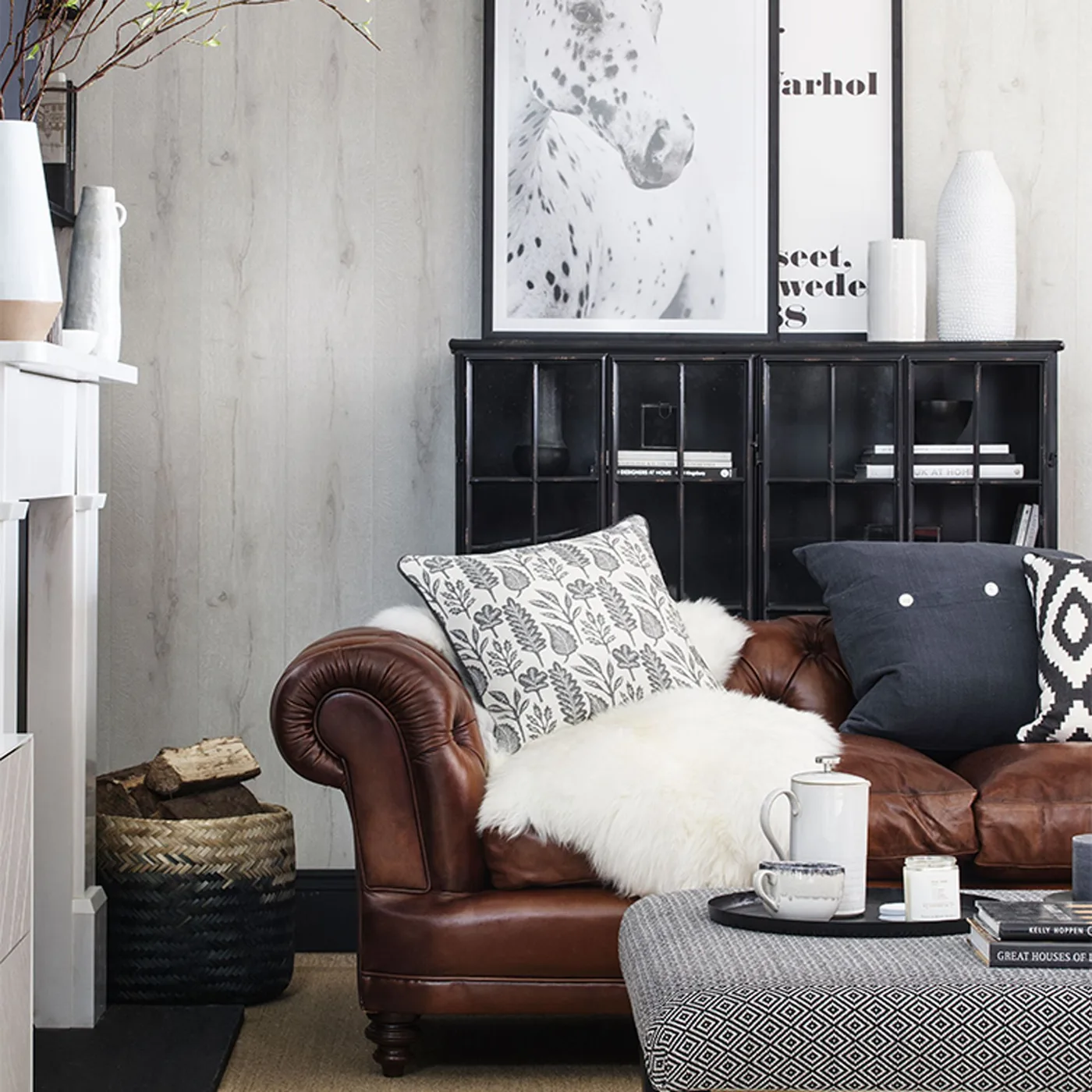 Brown rolled-arm couch with pale botanical pillow, dark buttoned pillow and black-and-white geometric pillow.