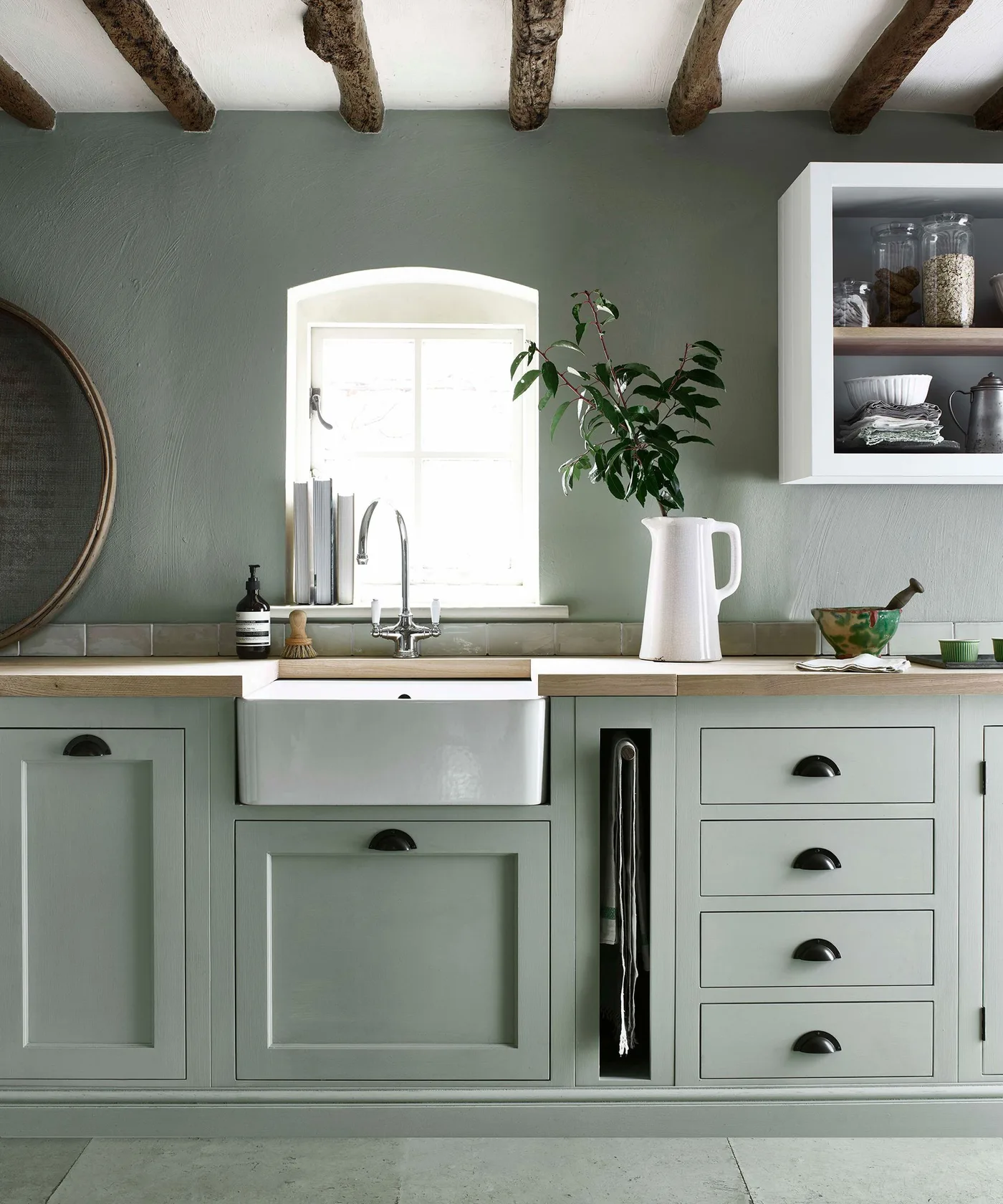 Country kitchen sink corner with green framed cabinets and drawers, white apron-front basin, pale wood-toned counter, small arched window and exposed beams.