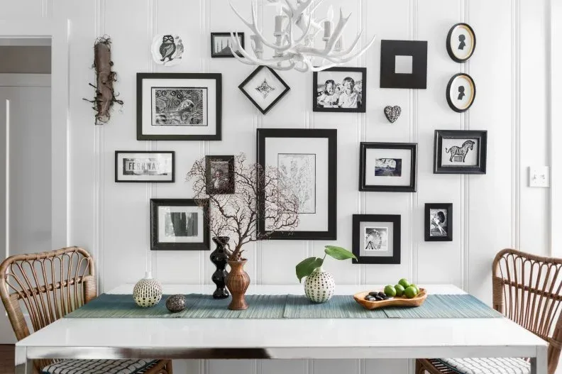 Black-and-white gallery with rectangular, oval, and diamond-shaped frames above a white dining table, wooden chairs, green runner, and pale antler-style chandelier.