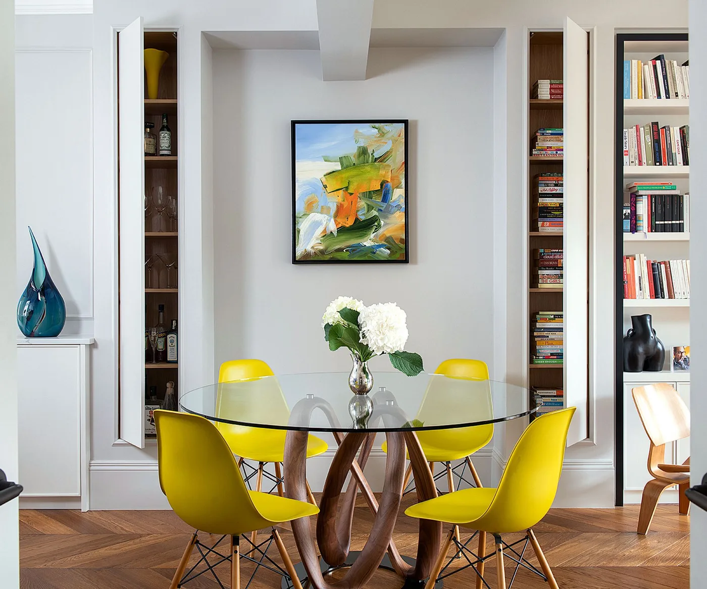 Tall narrow cupboards with open doors beside a recessed picture and a glass table with yellow chairs