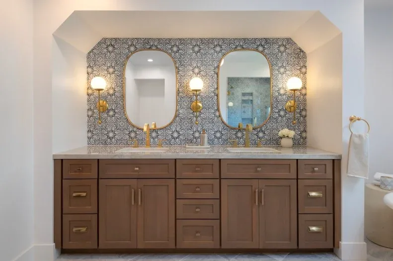 A wood double vanity with pale counter and warm-toned taps sits beneath two rounded mirrors and three globe wall lights beside a patterned blue-gray wall