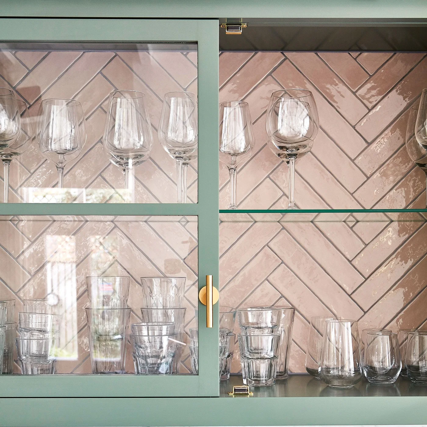 Pale pink herringbone tiles form the backing of a green glass-front cabinet holding drinking glasses