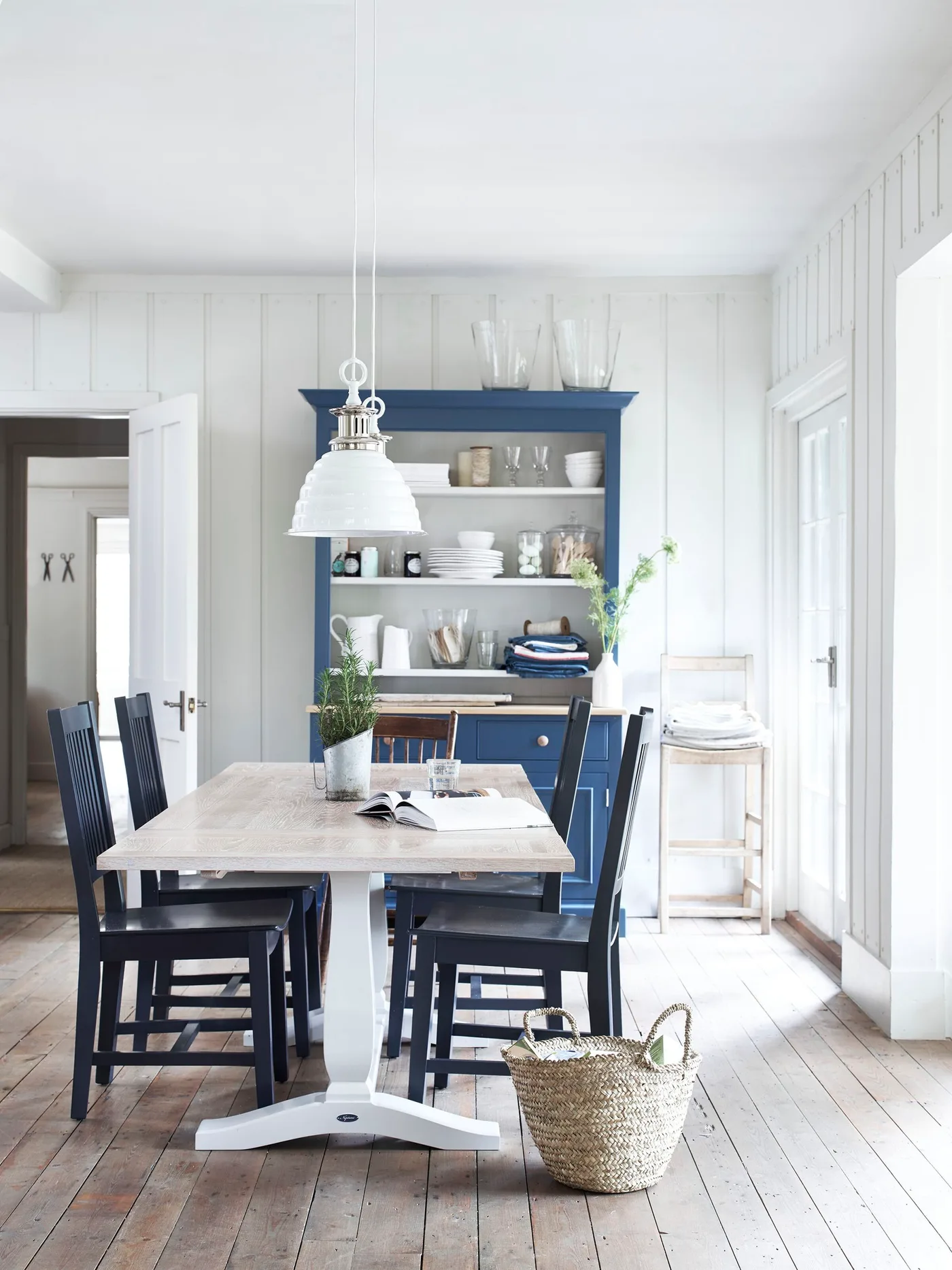 Pale paneled dining room with a light wooden table, blue chairs, blue dresser displaying dishes and a white pendant light.