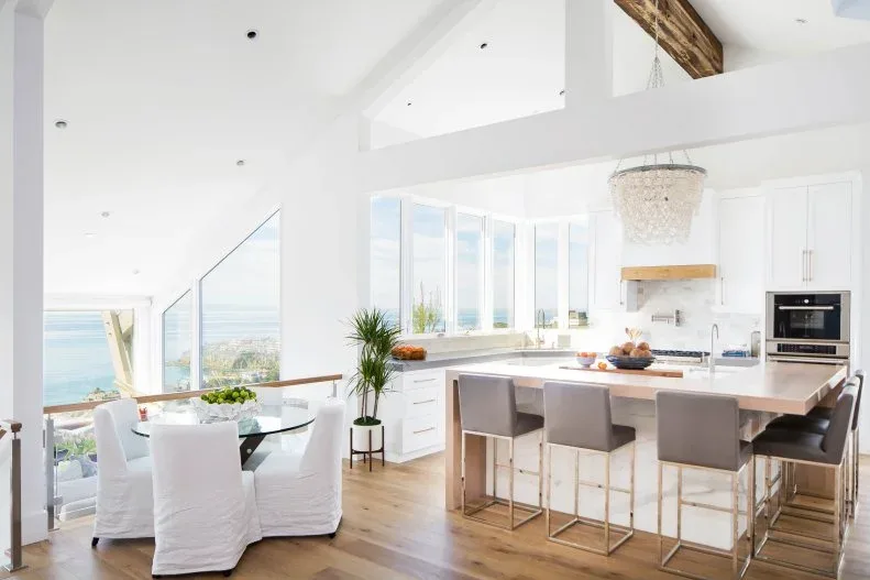 Open coastal kitchen with island stools and separate round dining table surrounded by white covered chairs beside windows.