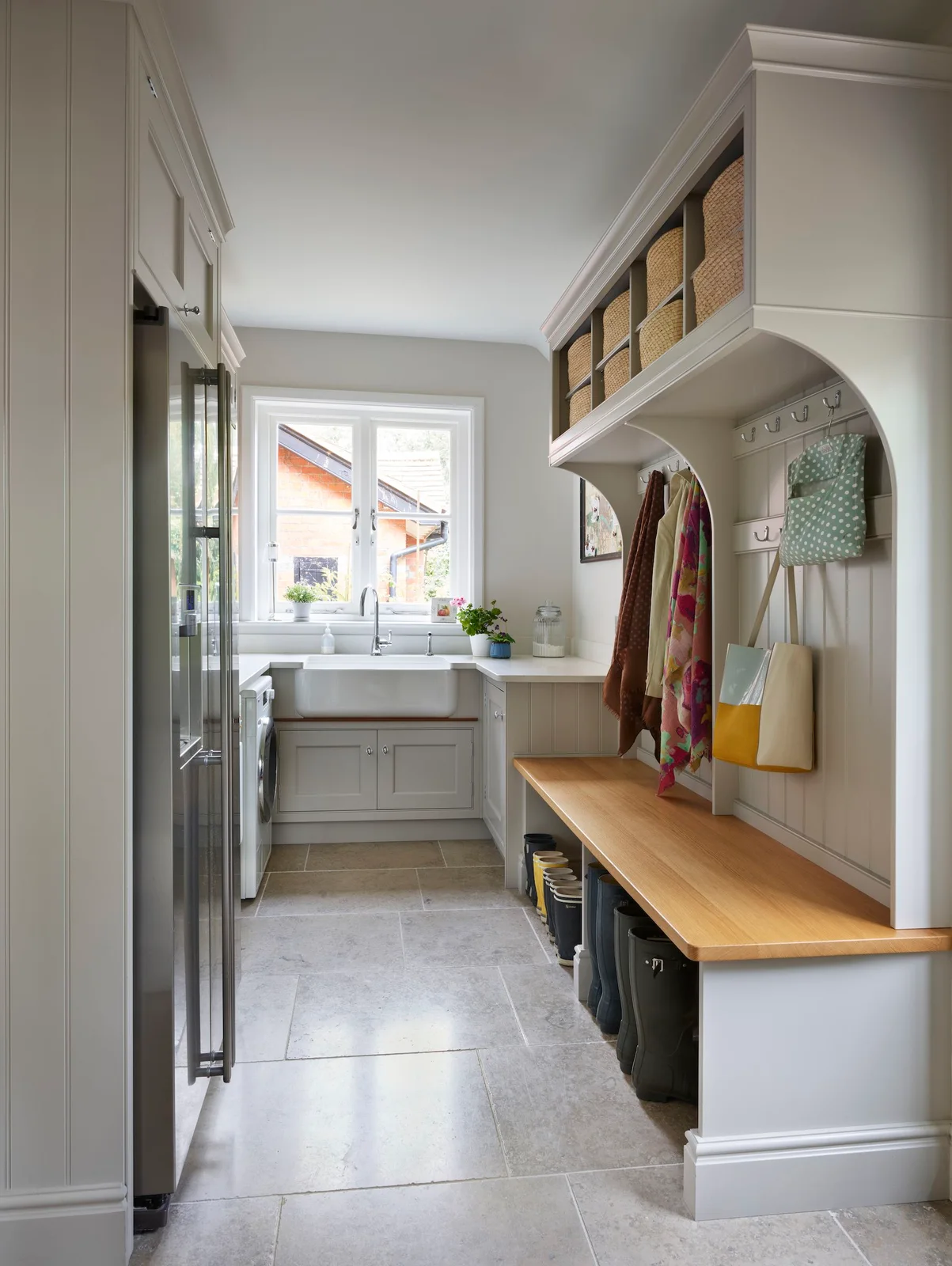 A wood-topped entry bench with coat hooks and basket cubbies stands opposite pale cupboards beside a white sink under a window and large pale floor tiles