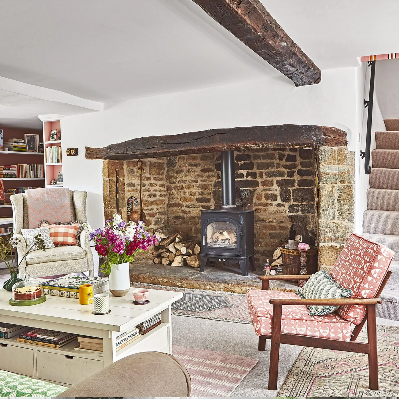 Broad stone inglenook with dark stove, wood beam and two differently patterned chairs around pale table