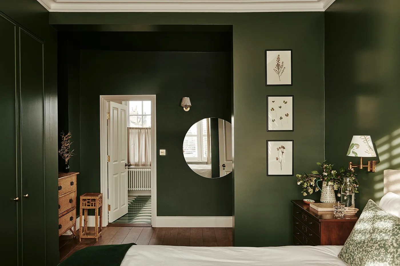 Deep green bedroom walls and cupboards beside pale bedding, botanical pictures and a round mirror