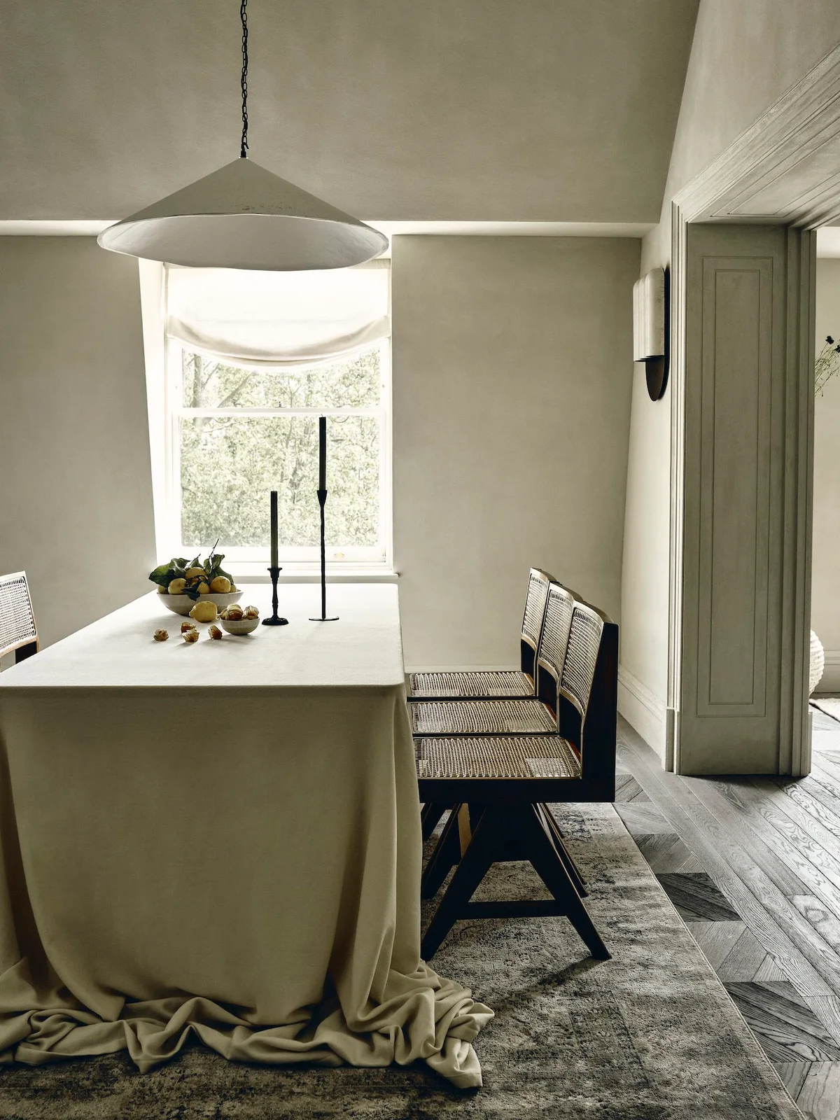 A muted green dining room has a long pale draped tablecloth, dark cane-backed chairs, a wide cone pendant and a softly gathered window shade