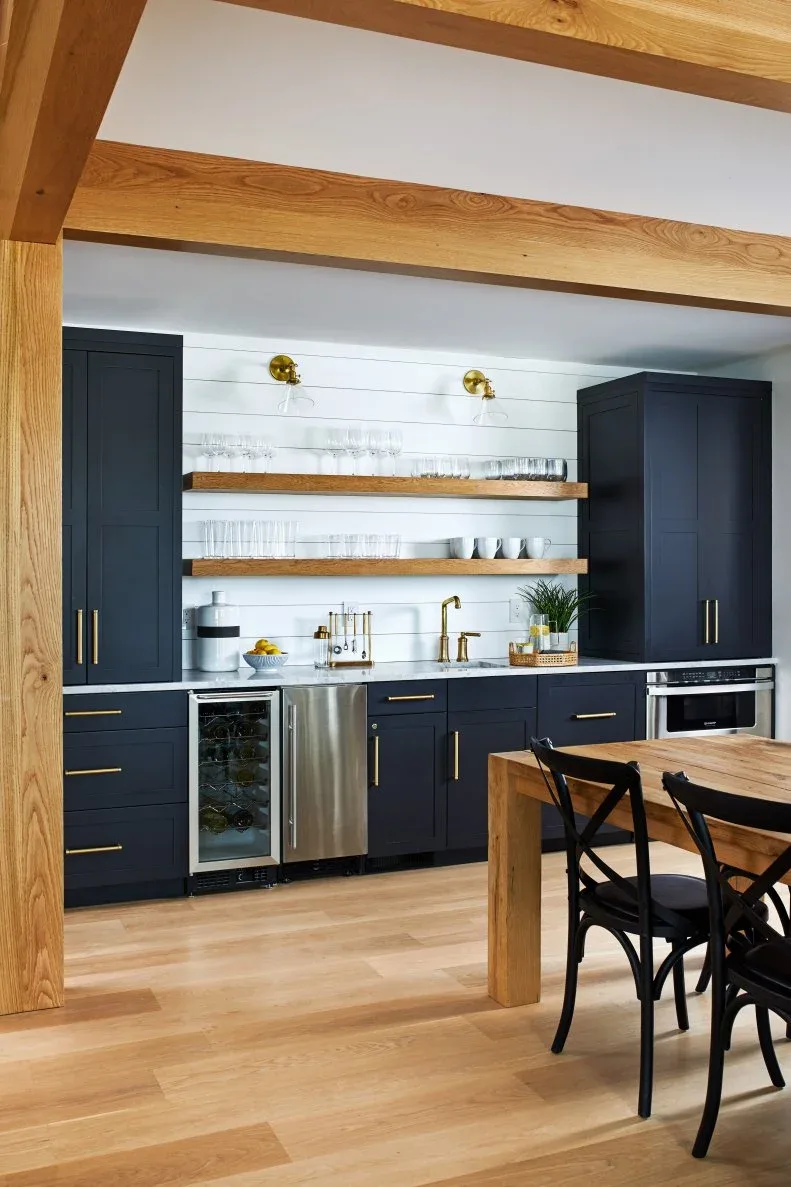 Two wood-toned floating shelves with clear glasses and white cups between dark cabinets beside a wooden dining table