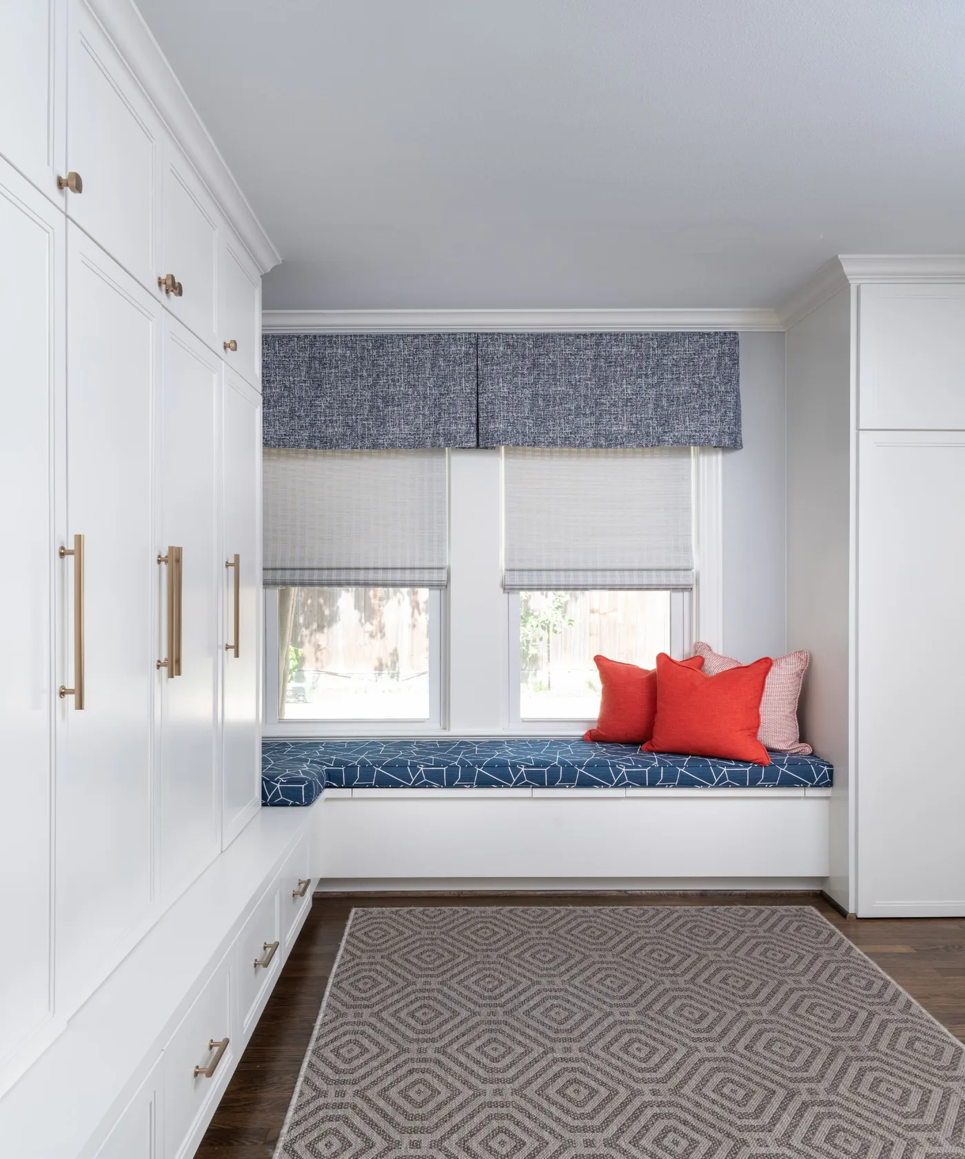 Pale entry storage room with tall white cupboards, long window bench, blue patterned cushion, red pillows and patterned pale rug