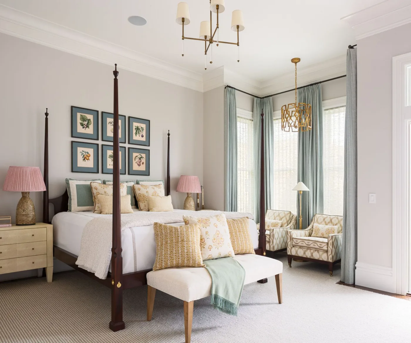 Country bedroom with a dark four-poster bed, six framed botanical pictures, pale bedding, pink bedside lamps and patterned chairs near long blue curtains.