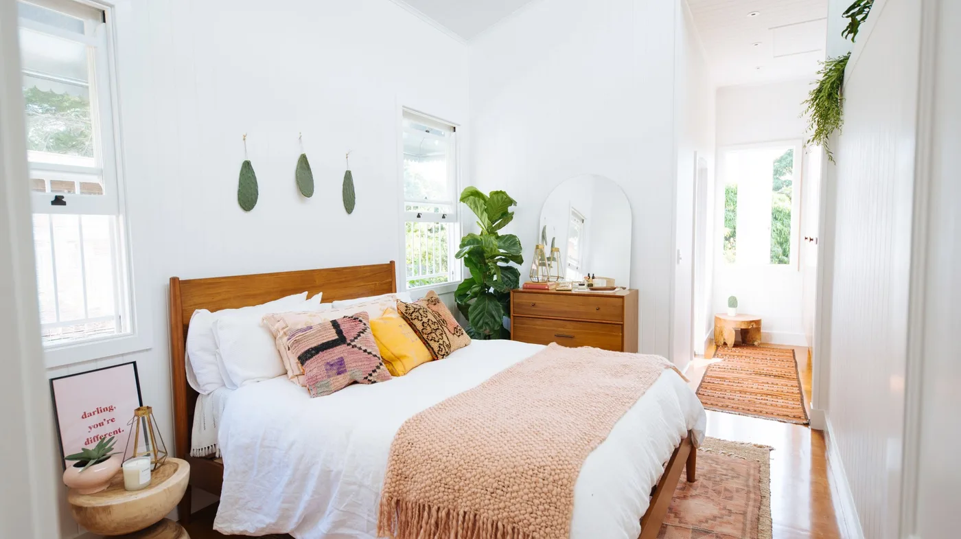 Bright bedroom with wooden bed and dresser, white bedding, pink throw, patterned cushions, small round bedside table and green plant.