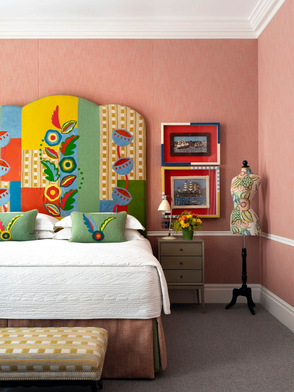 A tall rounded patchwork headboard combines colorful prints behind white bedding beside framed pictures and a yellow checked bench