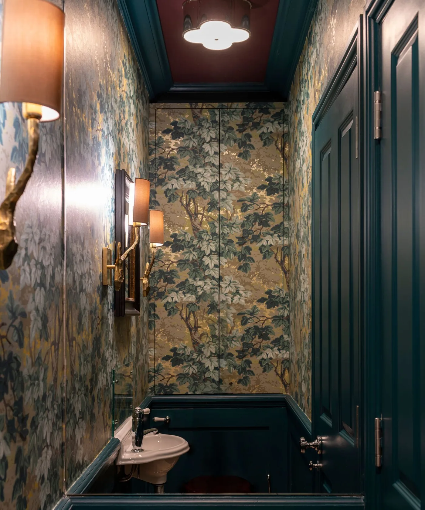 Narrow powder room with botanical wallpaper, deep green paneled door and lower walls, small pale basin, warm shaded lights and reddish ceiling.