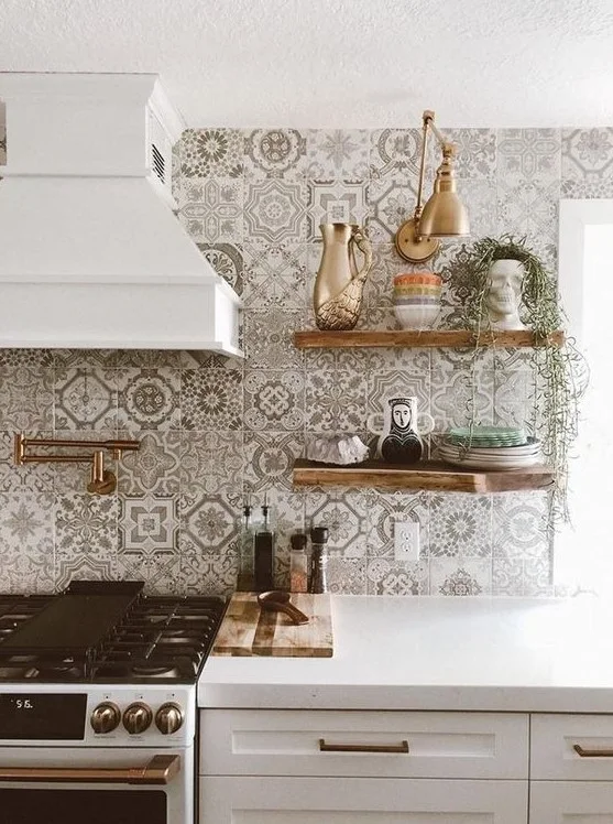 Pale patterned backsplash tiles surround a white hood beside wood-toned shelves, warm-colored wall light, plates and white base cabinets