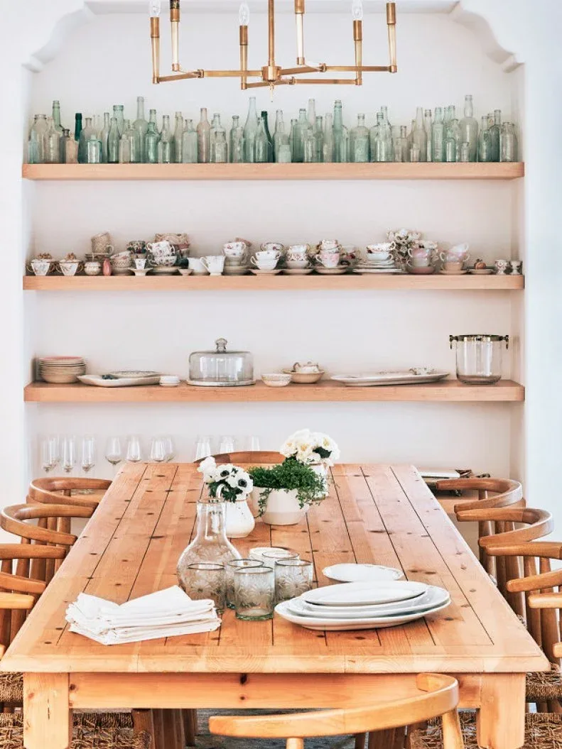 Three long wood shelves with pale green bottles, cups and serving pieces above a wood dining table