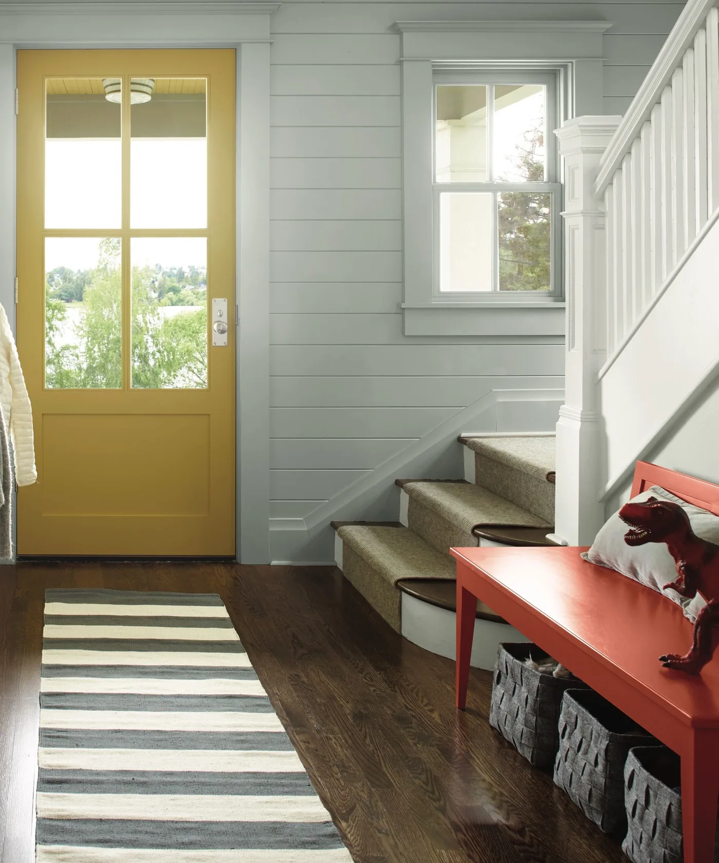 Entry with yellow glazed door, pale horizontal paneling, red bench, lower dark baskets, striped runner and white staircase