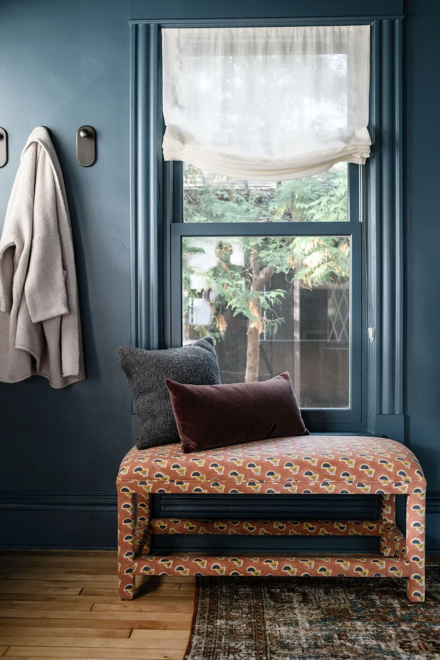 Blue mudroom nook with patterned upholstered bench, pale window shade, dark cushion and coat hook.