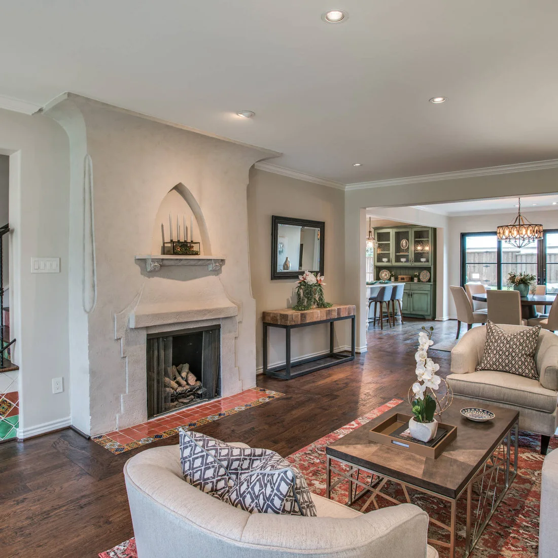 A sculptural pale fireplace with an arched niche stands beside patterned hearth tiles, pale living-room seats and a printed rug with a dining area beyond