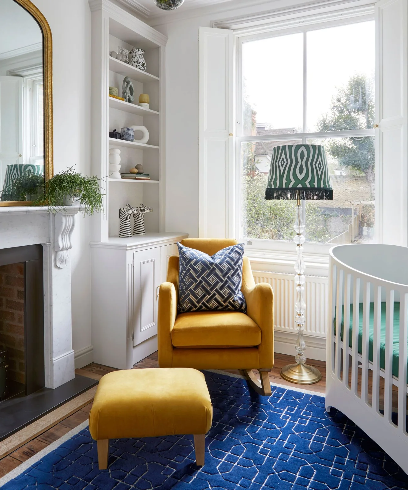 A mustard adult chair and matching footstool sit on a blue geometric rug beside a green patterned lampshade, white shelving and a curved white crib