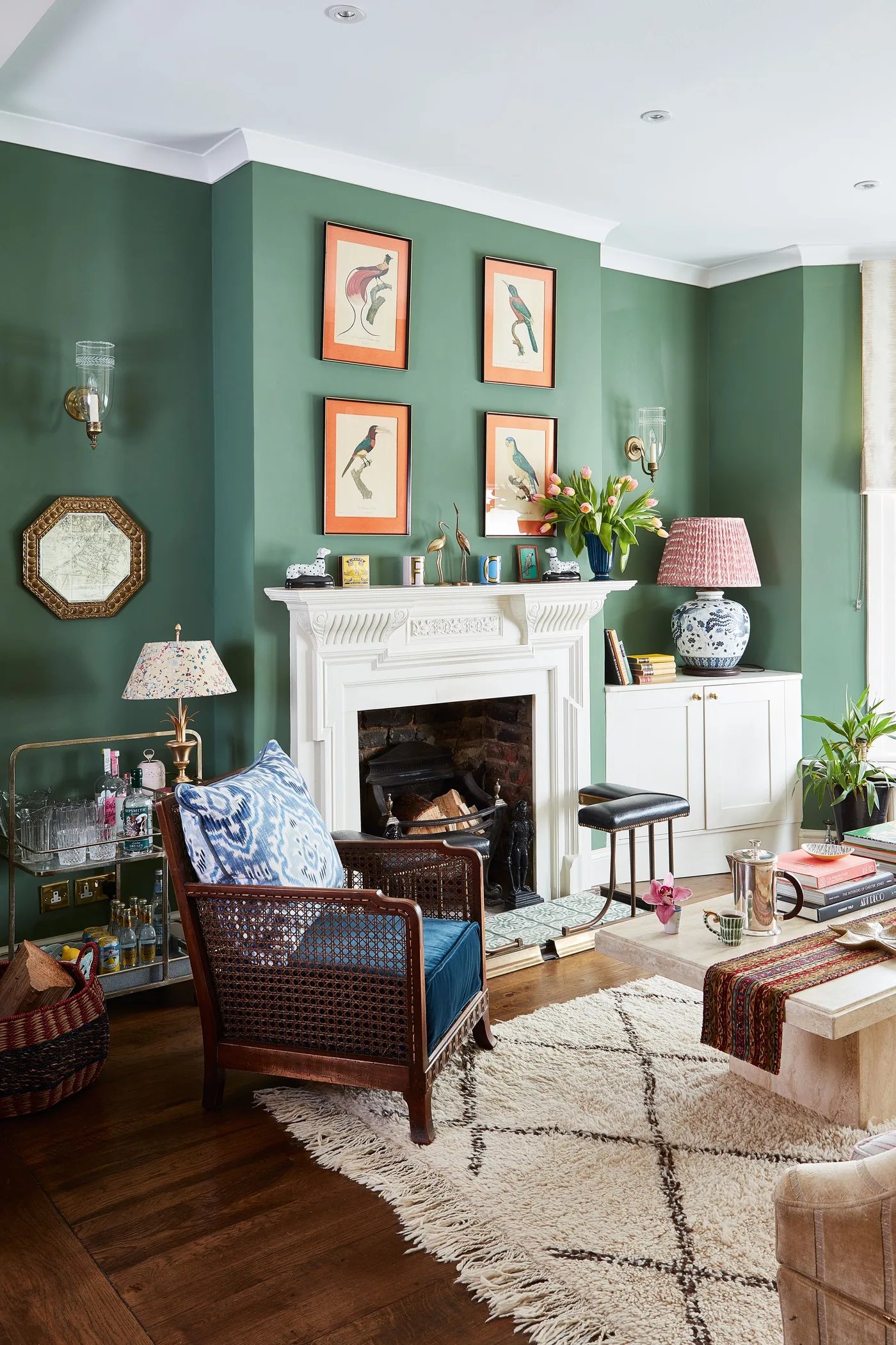 Green sitting room with four orange-mounted bird pictures above white mantel, blue chair and patterned lamps